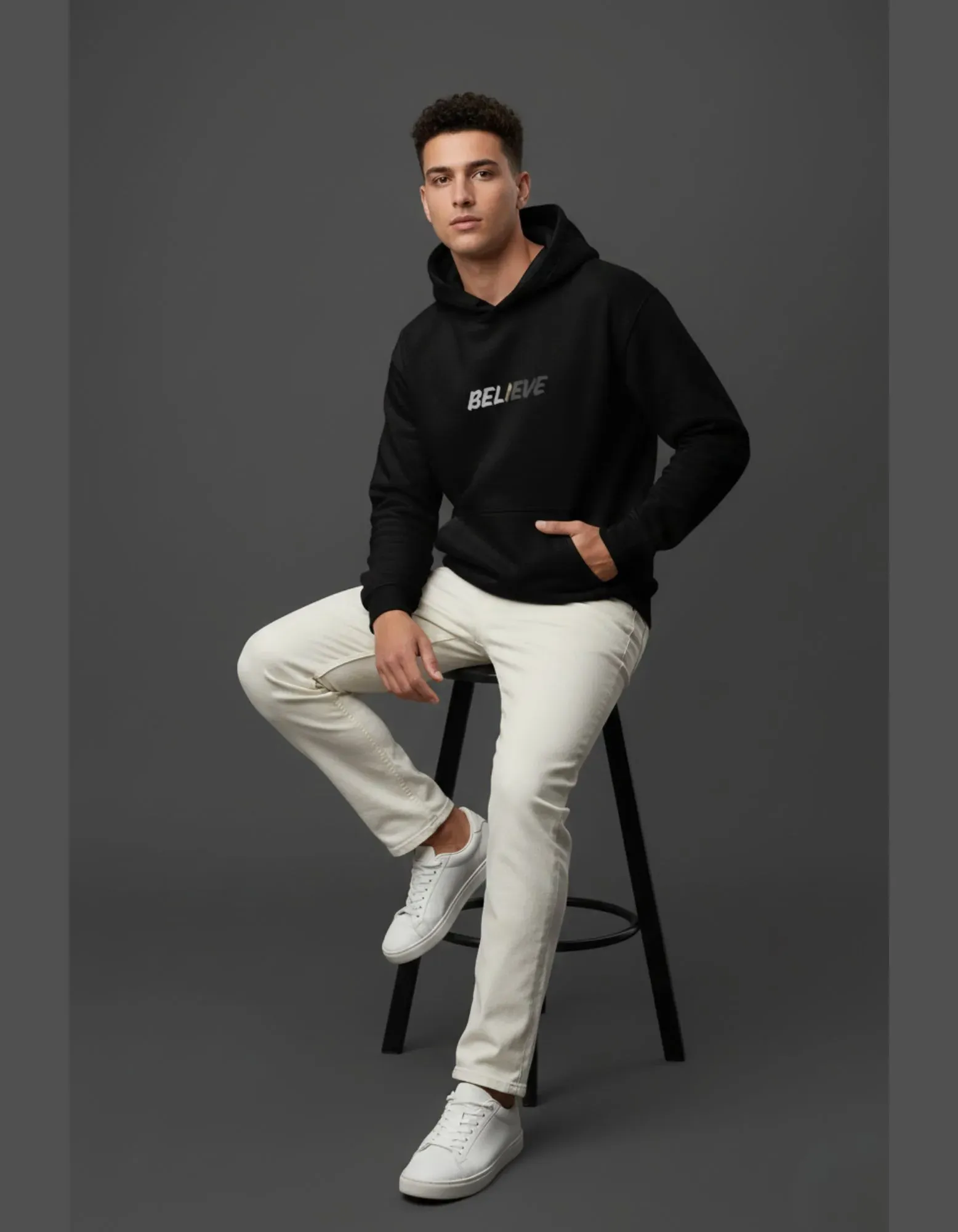 Man wearing a black hoodie with 'BELIEVE' text, white pants, and white sneakers on a gray background